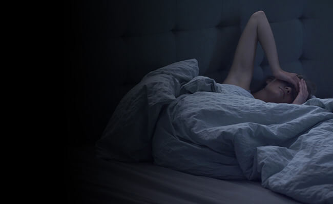Pessoa deitada na cama enrolada nos cobertores com as mãos sobre os olhos