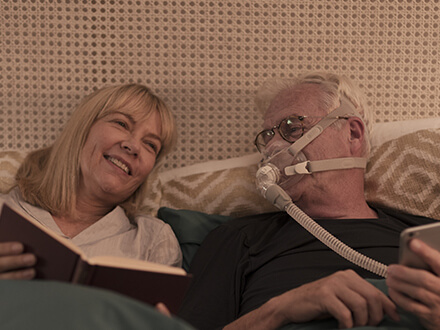 Paciente dorme ao lado da mulher enquanto beneficia da terapia usando a máscara facial minimalista da ResMed AirFit F30
