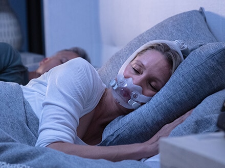 Casal dorme numa cama. Mulher utiliza uma máscara facial AirFit F30i que permite total liberdade de movimentos.