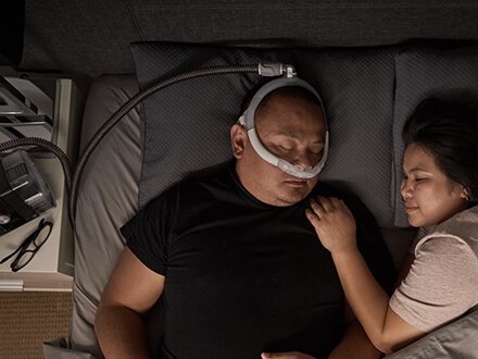 Casal dorme numa cama. O marido utiliza uma máscara de almofadas nasais AirFit P30i da ResMed para terapia.
