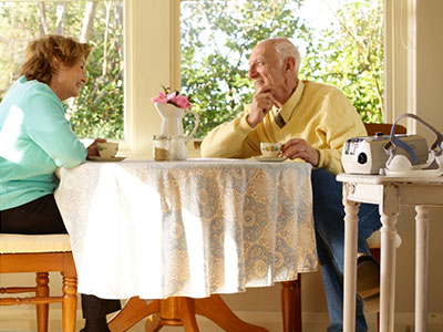 Casal de idosos sentado a uma mesa conversa. Dispositivo médico encontra-se numa mesa, ao lado.