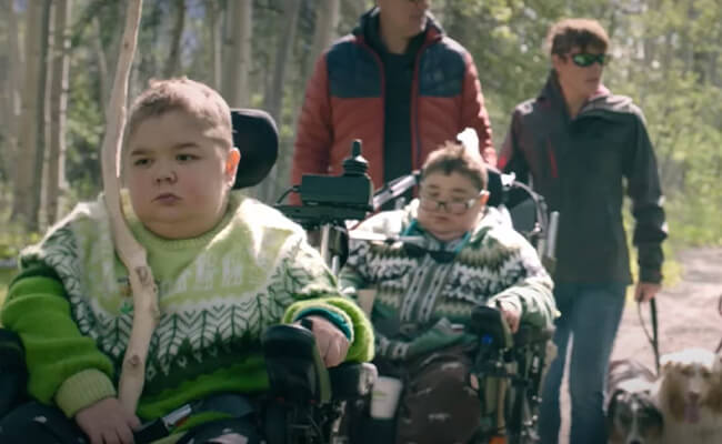 Duas crianças com doença neuromuscular em cadeira de rodas acompanhadas pelos seus pais no campo