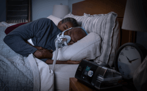 Casal dorme numa cama. Marido usa máscara de ResMed conectada a um dispositivo AirSense10.
