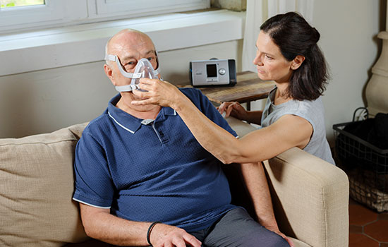 Paciente encontra-se sentado no sofá enquanto mulher lhe coloca uma máscara de terapia. O dispositivo encontra-se na imagem