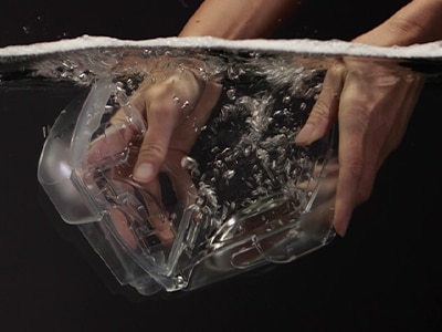 um par de mãos mergulhando uma câmara de humidificador de dispositivo CPAP ResMed em água contra um fundo preto