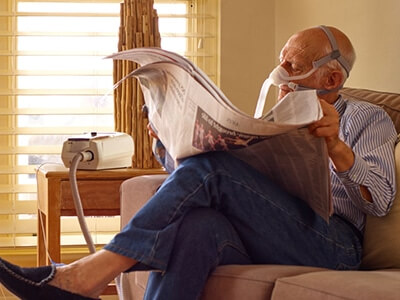 Homem de idade sentado a um sofá lê um jornal conectado a um dispositivo de terapia