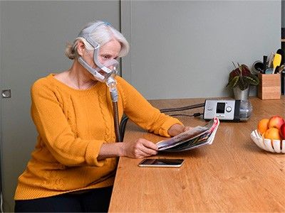 Senhora de idade sentada a uma mesa, conectada a um dispositivo e a realizar a sua terapia enquanto lê uma revista