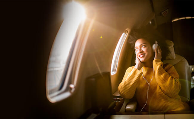 Mulher de phones nos ouvidos viaja de avião no lugar da janela, enquanto sol entra pela mesma