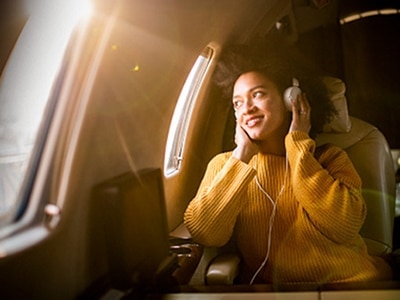 Uma jovem mulher a olhar pela janela de um avião enquanto usa auscultadores, com a luz do sol a entrar pela janela.