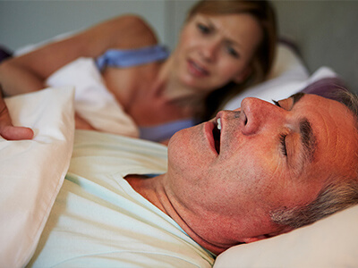 Casal está deitado numa cama. Homem dorme de boca aberta enquanto mulher olha indignada, incapaz de dormir